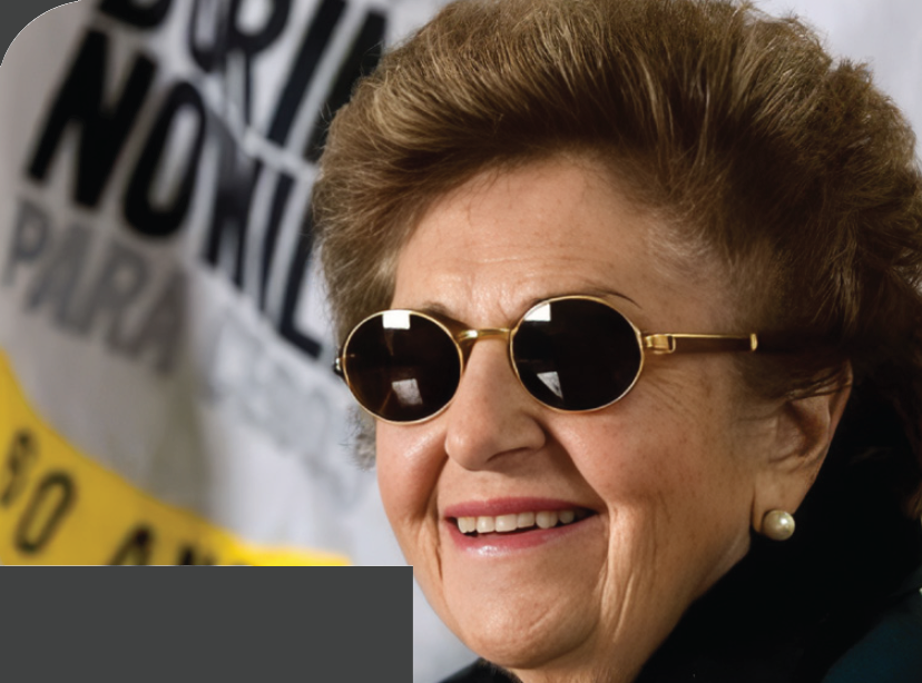 Imagem: Fotografia. Dorina Nowill, uma mulher com cabelo curto castanho, usa óculos escuros com armação dourada, brincos de pérola e blusa com gola preta. Ela sorri. Ao fundo, há um cartaz com o texto: DORINA NOWILL PARA CEGOS, 50 ANOS. Fim da imagem.