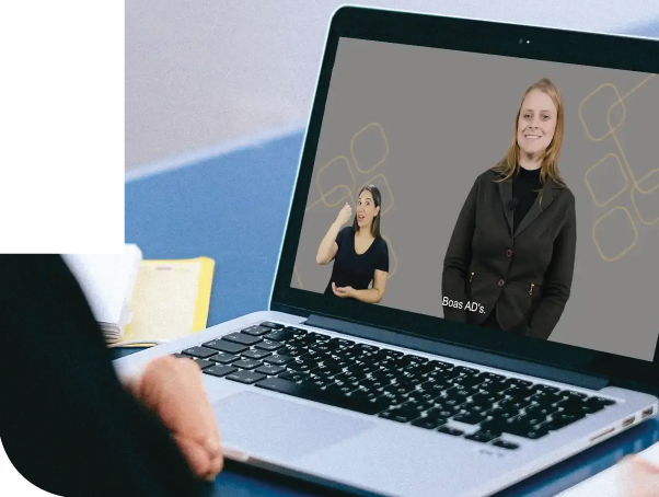 Imagem: Fotografia. Destaque para o torso de uma pessoa de costas sentada à mesa com a mão próxima ao teclado de um notebook. Na tela do notebook há um vídeo com uma mulher de cabelos loiros, usa blusa e casaco preto e uma mulher com cabelos pretos, usa blusa preta e sinaliza libras.  Fim da imagem.