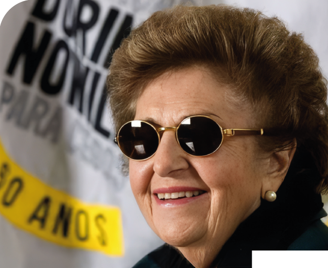 Imagem: Fotografia. Dorina Nowill, usa óculos escuros com armação dourada, brincos de pérola e blusa com gola preta. Ela sorri. Ao fundo, há um cartaz com o texto: DORINA NOWILL PARA CEGOS, 50 ANOS. Fim da imagem.