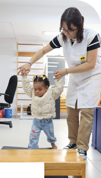 Imagem: Fotografia. À direita, uma mulher de cabelos castanhos e curtos, usa óculos, jaleco branco sobre camiseta preta, calça marrom e tênis preto, segura as duas mãos de uma menina de cabelos presos com laços amarelos, veste jaqueta amarela e calça azul, caminha sobre um colchonete preto, em frente de uma plataforma de madeira. Ao fundo, há uma cadeira preta, escada de madeira e lixeiras. Fim da imagem.