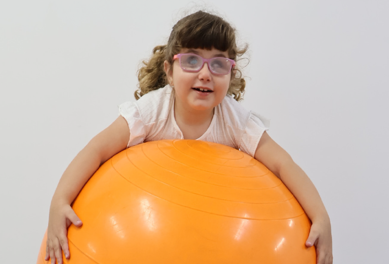 Imagem: Fotografia. Menina com cabelos castanhos cacheados presos para trás usa óculos com armação rosa e blusa branca, apoia o peito e os braços sobre uma bola grande laranja com linhas circulares em relevo; as mãos seguram as laterais da bola. Fim da imagem.
