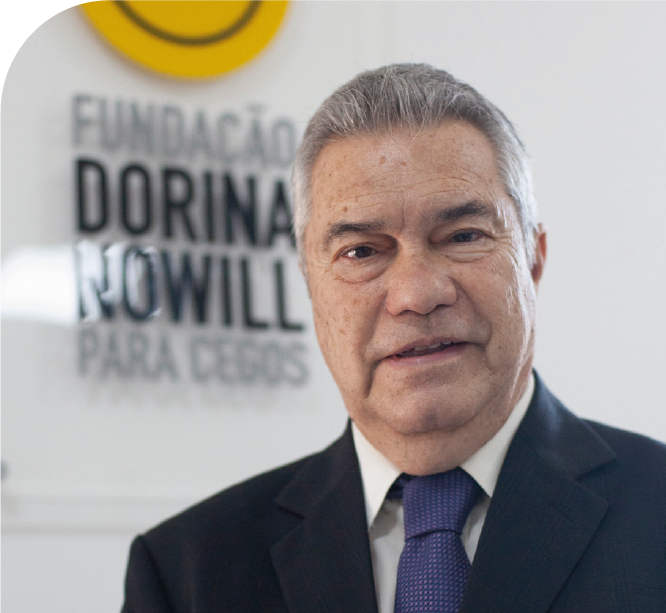 Imagem: Fotografia. Homem de cabelos curtos grisalhos usa terno preto, camisa branca e gravata azul, sorri. Ao fundo, à esquerda, há um painel desfocado com o logotipo da Fundação Dorina Nowill para Cegos. Fim da imagem.