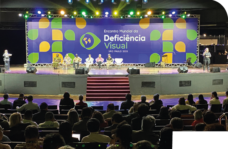 Imagem: Fotografia. Um palco amplo com um grande painel azul ao fundo com formas geométricas em verde e amarelo, o texto ‘Encontro Mundial da Deficiência Visual; SÃO PAULO, 2025’ e a ilustração da silhueta da anatomia de um olho com um globo terrestre. Sobre o palco, à esquerda, uma pessoa está em pé atrás de um púlpito; ao centro, várias pessoas estão sentadas em cadeiras alinhadas; à direita, uma pessoa está em pé sinalizando em libras. Na parte superior, há refletores com luzes coloridas em uma estrutura metálica. Na parte inferior, uma escadaria central com degraus vermelhos. Em frente ao palco, há várias fileiras de cadeiras ocupadas por uma plateia, com pessoas vistas de costas. Fim da imagem.