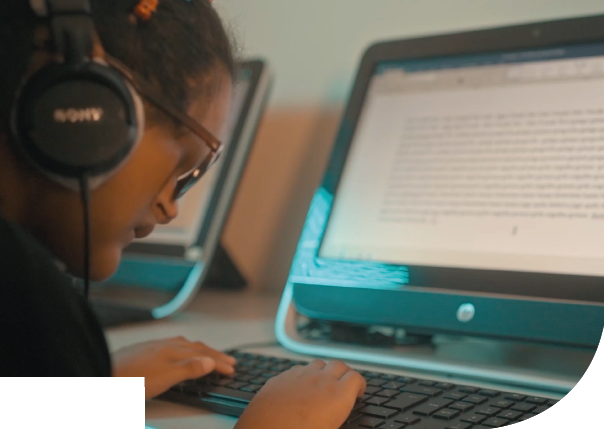Imagem: Fotografia. Destaque para o torso de uma pessoa com cabelos pretos presos, usa óculos escuros e fones de ouvido, está sentada à mesa com as mãos no teclado de um computador. O monitor exibe texto. Fim da imagem.