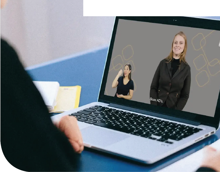Imagem: Fotografia. Destaque para o torso de uma pessoa de costas sentada à mesa com a mão próxima ao teclado de um notebook. Na tela do notebook há um vídeo com uma mulher de cabelos loiros, usa blusa e casaco preto e uma mulher com cabelos pretos, usa blusa preta e sinaliza libras. Fim da imagem.