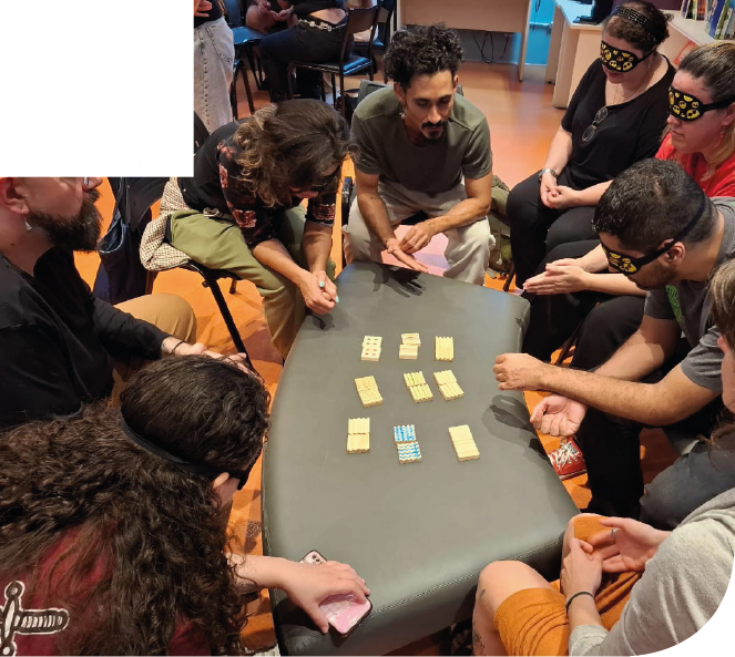 Imagem: Fotografia. Oito pessoas estão sentadas em círculo ao redor de uma mesa retangular cinza, com várias peças retangulares pequenas organizadas em grupos no centro e uma peça com listras azuis e brancas próxima à parte inferior. A maioria das pessoas está usando vendas pretas e amarelas sobre os olhos. Ao fundo, há cadeiras e pessoas sentadas, com piso em tom laranja e móveis brancos. Fim da imagem.