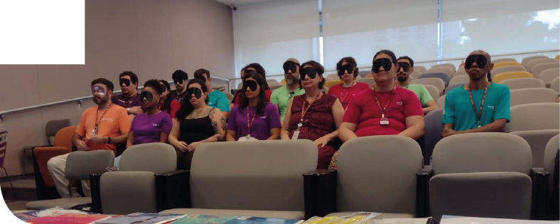 Imagem: Fotografia. Em um auditório com poltronas estofadas cinza organizadas em fileiras, diversas pessoas estão sentadas, voltadas para a frente, usando vendas pretas sobre os olhos e algumas usam crachás com cordão no pescoço. À frente, há uma mesa com papéis coloridos. Ao fundo, há janelas com persianas brancas. Fim da imagem.