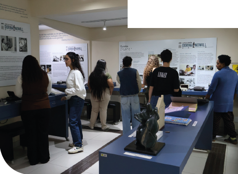 Imagem: Fotografia. Sala de exposição com paredes bege cobertas por painéis informativos com textos e fotos. Encostadas nas paredes e à frente, há mesas retangulares, compridas e azuis com objetos expostos. Diversas pessoas estão de pé e observam os painéis e os objetos. O piso é branco com piso tátil escuro, e o teto possui iluminação com trilhos e spots direcionados. Fim da imagem.