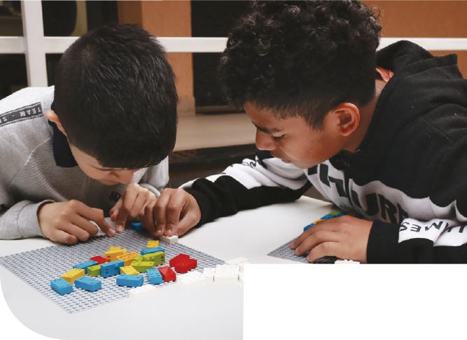 Imagem: Fotografia. Dois meninos estão inclinados sobre uma mesa branca e montam peças de lego Braille Bricks sobre uma base cinza. À esquerda, menino de cabelos curtos e pretos veste blusa cinza com gola escura e mantém a cabeça baixa, com as mãos próximas à base. À direita, menino de cabelos crespos e pretos veste moletom preto com detalhes brancos e mantém o rosto voltado para a montagem, com a mão direita sobre uma peça. Sobre a base, há peças coloridas espalhadas. Fim da imagem.