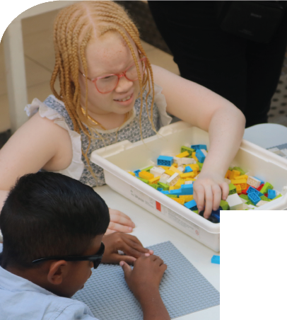 Imagem: Fotografia. Duas crianças estão sentadas à mesa e manipulam peças coloridas de Lego Braille Bricks. Ao centro, menina de cabelos longos e loiros em tranças usa óculos com armação vermelha e blusa sem mangas com babados, está com a mão esquerda dentro de uma caixa plástica branca com peças coloridas. À esquerda, menino de cabelos curtos e pretos usa óculos escuros e camiseta azul, apoia os antebraços sobre a mesa e pressiona uma peça sobre uma base quadrada cinza com pinos. Fim da imagem.