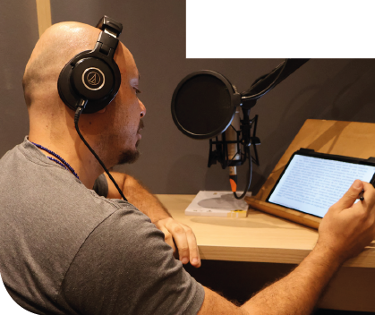 Imagem: Fotografia. Homem careca usa fones de ouvido pretos e camiseta cinza, está sentado à esquerda com os braços apoiados sobre uma mesa de madeira. Com a mão direita segura uma caneta sobre um tablet com texto. À frente, microfone preto com filtro circular preso a um suporte. Fim da imagem.