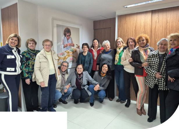 Imagem: Fotografia. Grupo com quatorze mulheres lado a lado sorrindo. Algumas estão em pé e outras agachadas. Atrás delas, há paredes cobertas por painéis de madeira e um quadro com a fotografia de Dorina Nowill de óculos escuros e blusa branca, bordando sobre um tecido estampado. Fim da imagem.
