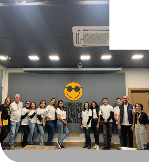 Imagem: Fotografia. Grupo de treze pessoas está em pé lado a lado. A maioria veste camiseta branca com logotipo azul. Na extremidades, duas mulheres vestem camiseta preta com o logotipo da Fundação Dorina Nowill para cegos. Atrás, painel cinza com o logotipo da Fundação Dorina. À direita, uma porta de madeira. Acima há um telão enrolado, luzes e ar-condicionado no teto. Fim da imagem.