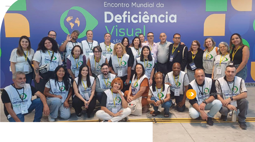 Imagem: Fotografia. Grupo de pessoas posa para foto. A maioria usa colete branco com o texto ‘Encontro Mundial da Deficiência Visual’ e crachás com cordões no pescoço. Algumas estão agachadas e outras em pé. Ao fundo, painel azul com formas arredondadas em verde e amarelo e texto: Encontro Mundial da Deficiência Visual; SÃO PAULO. Fim da imagem.