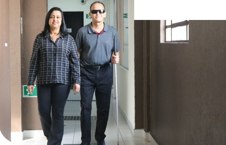 Imagem: Fotografia. Duas pessoas caminham lado a lado em um corredor interno. À esquerda, mulher de cabelos pretos e lisos, na altura dos ombros, veste camisa xadrez azul e branca e calça preta. À direita, homem de cabelos curtos e castanhos usa óculos escuros, camisa estampada cinza de manga curta e calça preta; ele segura uma bengala longa branca com a mão esquerda, posicionada à frente do corpo, enquanto segura o braços da mulher com a mão direita. À direita, janela com grades e vidro. O piso é branco com piso tátil sobre ele. Fim da imagem.