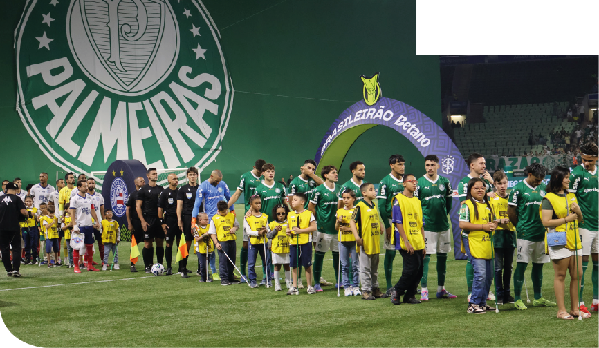 Imagem: Fotografia. Em um campo de futebol, fileiras de pessoas lado a lado. À esquerda, um grupo de homens veste uniformes brancos e verdes e há um homem de uniforme azul. Ao lado deles, há várias crianças usando coletes amarelos com o logotipo da Fundação Dorina Nowill para Cegos; algumas crianças seguram bengalas longas verdes ou brancas. Em seguida, há quatro homens de uniforme preto, dois deles seguram bandeiras amarela e laranja e ao lado, há uma bola de futebol. Atrás, grupo de homens de uniforme branco e preto e um homem de uniforme amarelo. Ao lado, há mais crianças de coletes amarelos. Ao fundo, uma arquibancada e um grande painel verde com um escudo circular e a palavra PALMEIRAS em branco. À direita, um arco roxo e verde com o texto BRASILEIRÃO Betano em branco. Fim da imagem.
