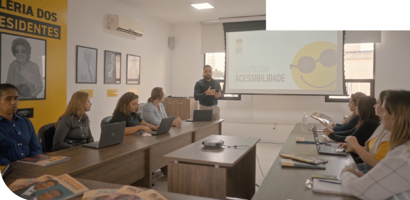 Imagem: Fotografia. Em uma sala de reunião, um homem está em pé ao fundo, gesticulando. Atrás dele há uma tela de projeção com a ilustração de um rosto sorridente usando óculos escuros e o texto SOLUÇÕES EM ACESSIBILIDADE. À frente, há diversas pessoas sentadas atrás de mesas marrons dispostas em formato retangular. Sobre as mesas, há notebooks e livros. Ao fundo, janelas com persianas brancas entreabertas, e na parede da esquerda, há uma parede amarela e branca com retratos em preto e branco e o texto GALERIA DOS PRESIDENTES. Fim da imagem.