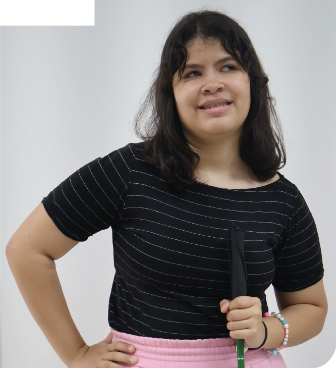 Imagem: Fotografia. Mulher de cabelos pretos na altura dos ombros, com franja, veste camiseta preta com listras finas em branco e calça rosa, usa pulseiras no pulso esquerdo, segura com a mão esquerda uma bengala verde na vertical, está com a mão direita na cintura e sorri. Fim da imagem.