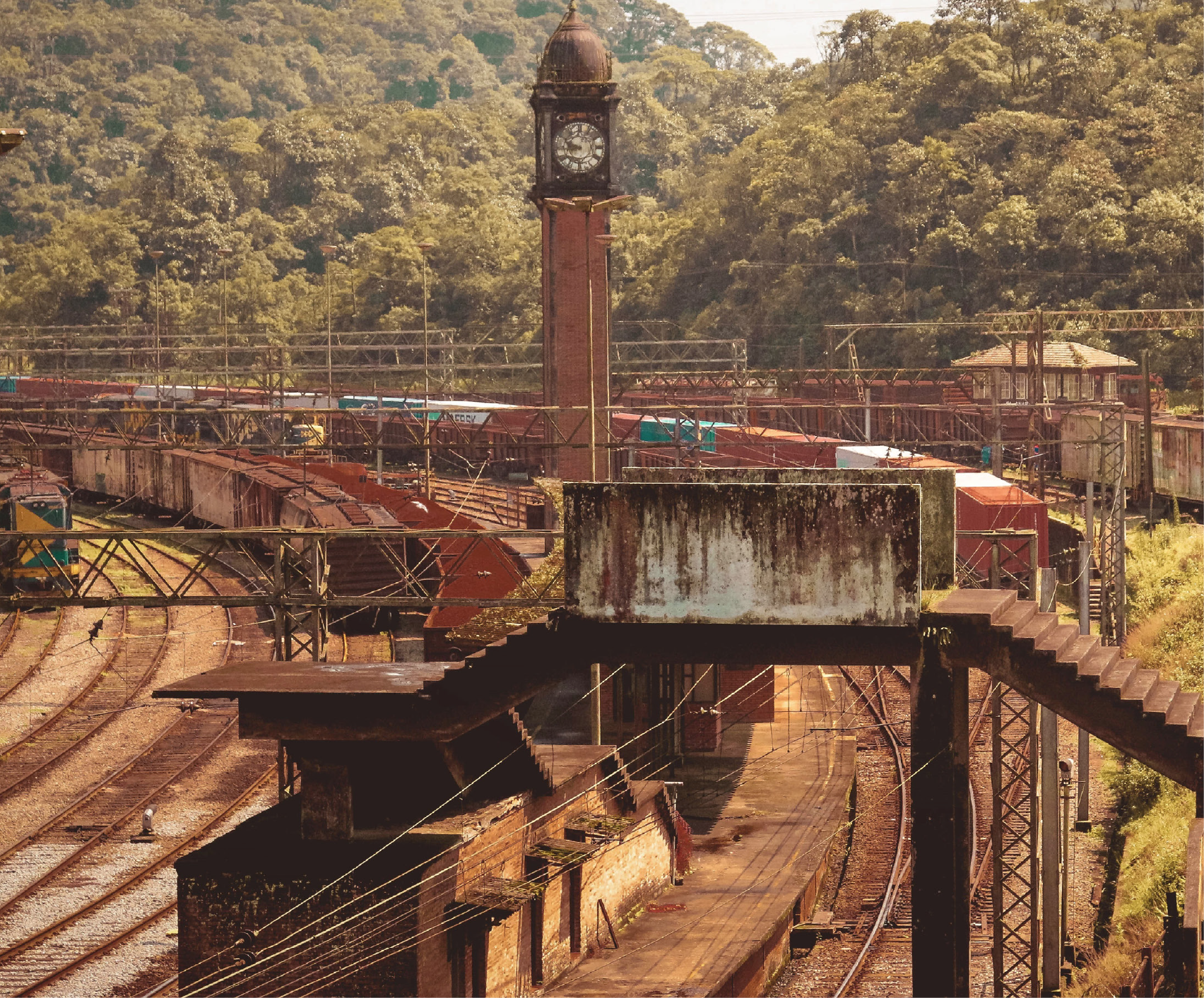 Imagem: Fotografia. Ao centro, uma torre alta de tijolos marrons com um relógio com número romanos e uma cúpula no topo. Abaixo e ao redor da torre, há uma área ferroviária com vários trilhos curvados, vagões e locomotivas em tons de vermelho e marrom, com alguns em azul e branco, e estruturas metálicas com vigas e passarelas. Na parte inferior, uma passarela elevada de concreto e metal atravessa os trilhos, com escadas nas duas extremidades. Ao fundo, uma encosta coberta por vegetação. Fim da imagem.