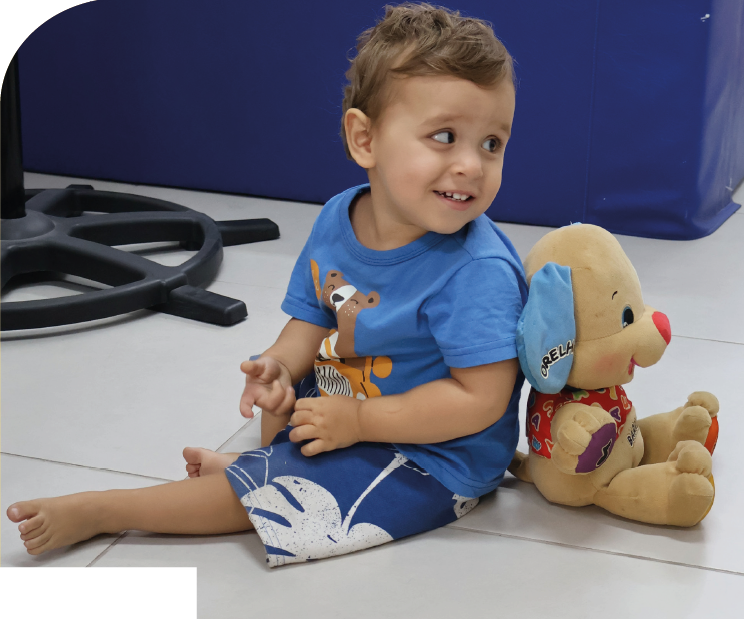 Imagem: Fotografia. Menino de cabelos curtos e castanhos usa camiseta azul com estampa e bermuda azul com desenhos brancos, está sentado no chão de piso branco, com uma perna estendida e outra dobrada, sorri com o rosto voltado para a direita. À direita, há um cachorro de pelúcia bege sentado encostado nas costas do menino, com orelha azul, nariz vermelho e roupa vermelha com estampa. À esquerda, há uma base preta de equipamento com formato circular. Fim da imagem.
