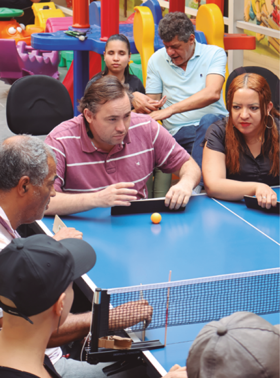 image: Photograph. People sitting around a tennis table. In the center, a man with short brown hair wears a pink polo shirt with white stripes and holds an adapted racket, consisting of a horizontal black rectangular support. Next to the adapted racket is a table tennis ball. On the right, a woman with long red hair wears a black blouse and holds the adapted racket. On the left, two men: one has short gray hair and is sitting on the side of the tennis table; the other wears a black cap and is sitting on the side of the table at the net. In the background, a girl with her eyes closed and a man holding her hands are sitting on chairs; behind them are playground toys. end of image.
