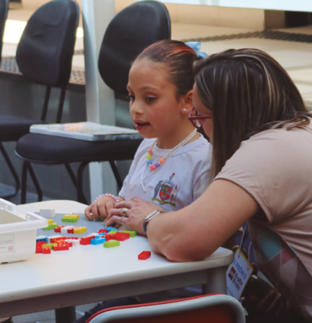 image: Photograph. A girl with brown hair tied in a ponytail wears a white blouse with a crest on her chest. She is sitting on a school desk chair with her hands on building blocks on the surface. Next to her, crouching, is a woman with brown hair, wearing glasses and a brown blouse. She has one hand on one of the girl's hands. In the background are black chairs. end of image.