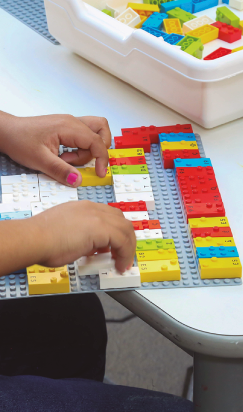 image: Photograph. Focus on a child's hands as she plays with colorful building blocks on a board. In the right corner, there is a white box with more colorful building blocks. end of image.