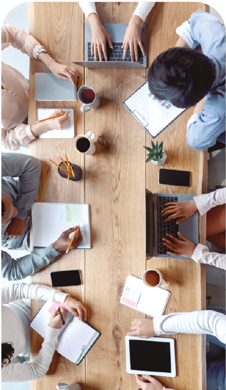 image: Photograph. Aerial view of a rectangular wooden table surrounded by people. On the table are mugs of coffee, notebooks, pencils, and tablets. end of image.