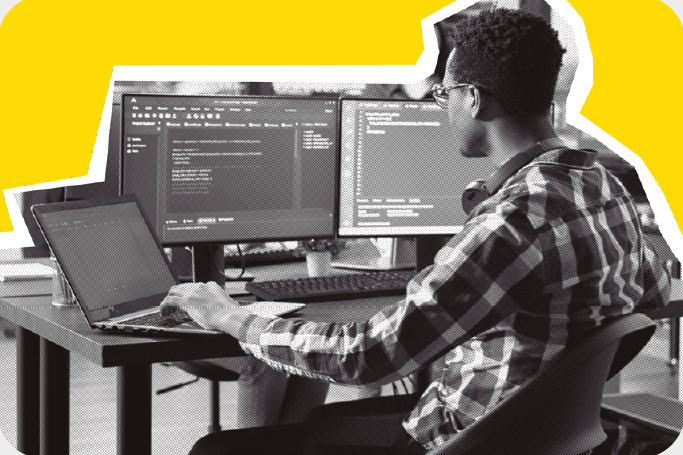 image: Black and white photograph. A man with short, curly hair, wearing glasses, a long-sleeved plaid shirt, pants, and headphones hanging around his neck. He is sitting in front of a desk, typing on a laptop on his left. On the right are two monitors positioned side by side, both displaying code on the screen, and a keyboard. end of image.