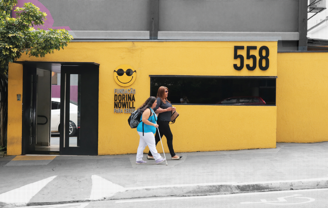 Imagem: Fotografia. Duas mulheres caminham na calçada, à frente de um prédio amarelo com o número "558" em preto na parte superior direita da fachada e o logotipo da FUNDAÇÃO DORINA NOWILL PARA CÉGOS. A mulher à esquerda possui cabelos pretos compridos, usa blusa azul, calça branca e tênis brancos, carrega uma mochila nas costas e segura uma bengala longa na mão. Ela está de braço dado com a mulher à direita. A mulher à direita possui cabelos longos castanhos compridos, usa óculos escuros, blusa cinza, calça preta e sapatilhas béges. Fim da imagem.