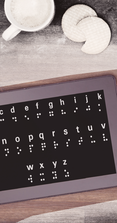 Imagem: Fotografia. Um tablet exibe o alfabeto de 'a' a 'z', acompanhado da respectiva representação em braille abaixo de cada letra. Acima, uma xícara branca com espuma na superfície e dois biscoitos. Fim da imagem.
