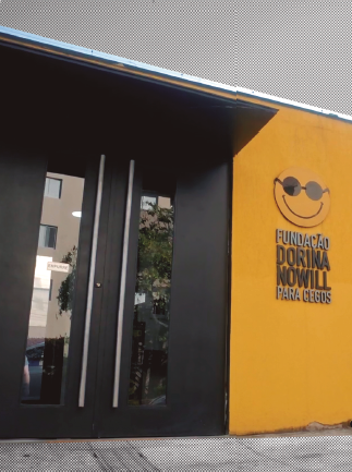 Imagem: Fotografia. Fachada de prédio com parede amarela à direita, onde há o logotipo da FUNDAÇÃO DORINA NOWILL PARA CEGOS. À esquerda, portas duplas pretas. Fim da imagem.