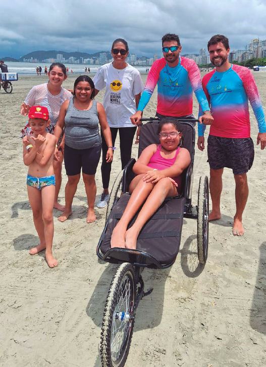 Imagem: Fotografia. Pessoas em uma praia. Ao centro, uma menina com cabelos pretos presos em um rabo de cavalo, usa óculos e maiô rosa. Ela está em uma cadeira de rodas adaptada, composta por três rodas e um suporte horizontal para as pernas. Atrás, segurando a cadeira, um homem com cabelos castanhos curtos e barba, usa óculos de sol e blusa térmica rosa e azul. À direita, outro homem com cabelos castanhos curtos e barba, usa blusa térmica rosa e azul e bermuda cinza. À esquerda, uma mulher com cabelos pretos presos em um rabo de cavalo, usa óculos escuros, camiseta branca com o logotipo da Fundação Dorina Nowill, légguing preta e tênis azuis. Ao lado dela, duas mulheres e uma criança. Fim da imagem.