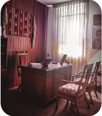 Imagem: Fotografia. Uma sala com parede de madeira à esquerda com um tapete vermelho com padrões geométricos pregado na parede. Ao centro, há uma mesa retangular de madeira com dois porta-retratos e um livro sobre ela. Atrás da mesa, há uma cadeira estofada com encosto alto e braços de madeira. À frente da mesa, duas cadeiras de madeira com assento almofadado estão lado a lado. No canto da parede à direita, parte de um quadro com retrato em preto-e-branco de Dorina Nowill. Fim da imagem.