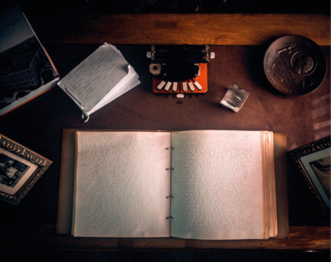 Imagem: Fotografia. Um livro aberto com escrita em braile nas páginas sobre uma mesa de madeira. Ao redor, uma máquina de escrever laranja, um prato decorativo redondo com alto relevo, um bloco de anotações, dois porta-retratos e um enfeite de vidro. Fim da imagem.