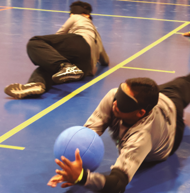 Imagem: Fotografia. Dois homens com cabelos curtos, usam vendas pretas e uniforme composto por blusa de manga comprida cinza, calça preta e tênis pretos. Eles estão deitados no chão de uma quadra esportiva azul, delimitada por linhas amarelas. O homem à direita segura uma bola azul entre as mãos. Fim da imagem.