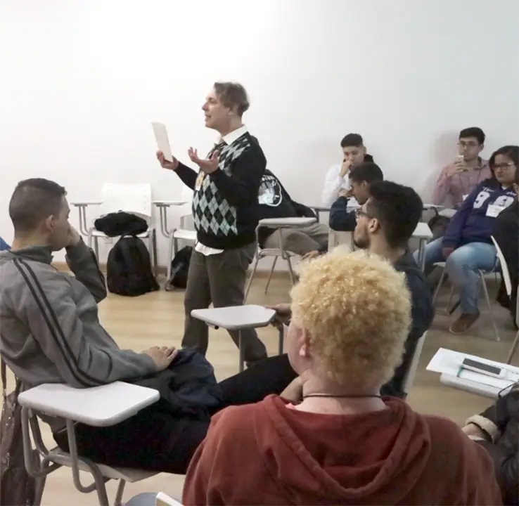 Foto de uma sala com pessoas sentadas em cadeiras assistindo o professor dando aula ao centro.
