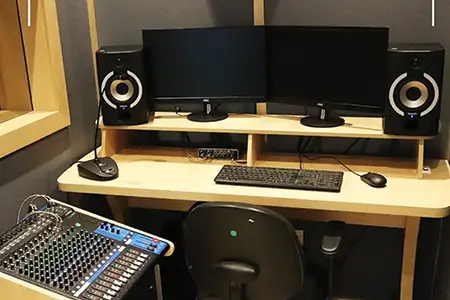 Foto de uma mesa dentro de um estúdio de áudio de paredes cinza. Sobre a mesa há duas caixas de som, dois monitores, uma placa de áudio, um microfone pequeno, mouse e teclado. Ao lado da mesa, à esquerda, há uma mesa de som sobre um suporte. Ela tem diversos botões e cabos conectados. Há ainda uma cadeira à direita da mesa de som. Fim da descrição.