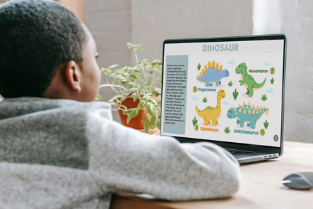 Foto de um menino em uma mesa com um notebook a frente. O menino está à esquerda, parcialmente de costas e usando um moletom cinza. À direita, o notebook está aberto e na tela há um livro digital com texto e diversos desenhos de dinossauros. Ao lado do notebook há ainda um vaso com planta. Fim da descrição.