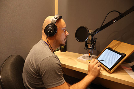 Foto de um narrador dentro de um estúdio de paredes cinza. Ele está à esquerda, sentado de perfil, e voltado para a direita onde há uma mesa com um apoio onde há um tablet aberto e também um pedestal com um microfone profissional. O narrador usa camiseta cinza e está com a mão direita segurando uma caneta digital próxima a tela do tablet. Fim da descrição.