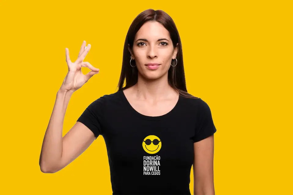 Foto de uma mulher, posicionada da cintura para cima, sobre fundo amarelo. Ela usa camiseta preta com o logo da Fundação Dorina e está com a mão direita levantada ao lado do rosto, fazendo um gesto com os dedos do meio e polegar juntos. Fim da descrição.