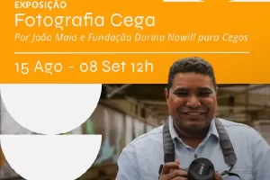 A imagem é um cartaz de divulgação de uma exposição chamada "Fotografia Cega". No lado esquerdo, há a foto de João Maia, que está sorridente e segura uma câmera fotográfica. Ele veste uma camisa azul clara e tem uma alça de câmera no pescoço. No lado direito, em um fundo laranja, estão as informações da exposição: - "Exposição Fotografia Cega" - "Por João Maia e Fundação Dorina Nowill para Cegos" - "15 Agosto a 08 Setembro, às 12h" - "Expressões Culturais" - "Unibes Cultural" Há também um selo indicando que a entrada é gratuita.