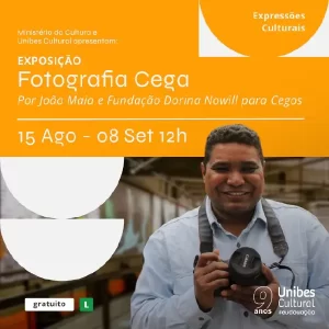 A imagem é um cartaz de divulgação de uma exposição chamada "Fotografia Cega". No lado esquerdo, há a foto de João Maia, que está sorridente e segura uma câmera fotográfica. Ele veste uma camisa azul clara e tem uma alça de câmera no pescoço. No lado direito, em um fundo laranja, estão as informações da exposição: - "Exposição Fotografia Cega" - "Por João Maia e Fundação Dorina Nowill para Cegos" - "15 Agosto a 08 Setembro, às 12h" - "Expressões Culturais" - "Unibes Cultural" Há também um selo indicando que a entrada é gratuita.