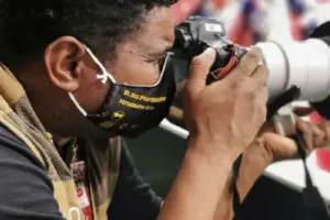 A imagem mostra o fotógrafo usando uma máscara preta e uma camiseta preta da Fundação Dorina Nowill para Cegos com um colete bege. Ele está segurando uma câmera com uma lente grande e branca, aparentemente tirando fotos em um evento esportivo. No fundo, há uma área desfocada com cores vermelhas e brancas, sugerindo que ele está em um ambiente movimentado, possivelmente uma competição.