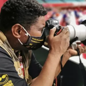 A imagem mostra o fotógrafo usando uma máscara preta e uma camiseta preta da Fundação Dorina Nowill para Cegos com um colete bege. Ele está segurando uma câmera com uma lente grande e branca, aparentemente tirando fotos em um evento esportivo. No fundo, há uma área desfocada com cores vermelhas e brancas, sugerindo que ele está em um ambiente movimentado, possivelmente uma competição.