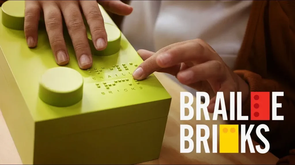 Uma pessoa toca em um grande tijolo verde amarelo com pontos em braile em relevo na parte superior. As palavras BRAILLE BRICKS aparecem ao lado do tijolo, com algumas letras substituídas por formas coloridas de tijolos.